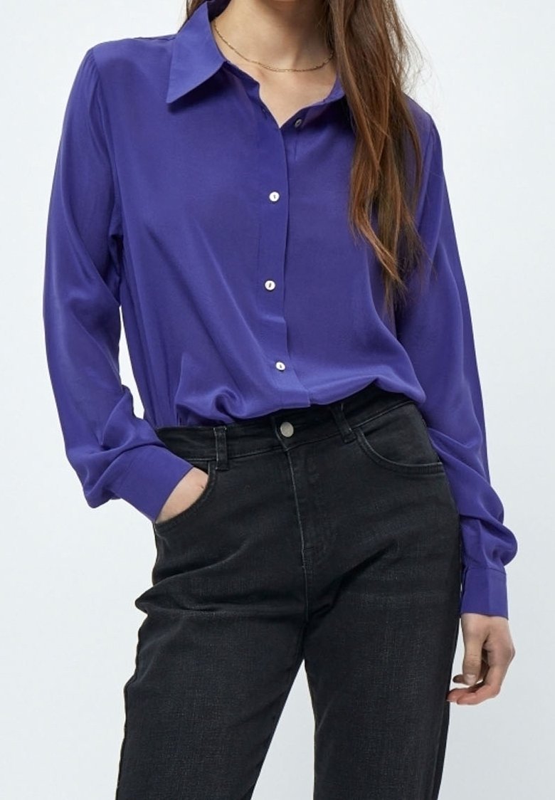 Femme portant une chemise violette à manches longues boutonnée, rentrée dans un jean noir taille haute, une main dans la poche, sur un fond clair.