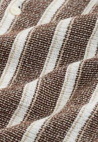 Gros plan sur un tissu tissé à rayures brunes et blanches texturé avec des coutures visibles et un petit bouton beige.