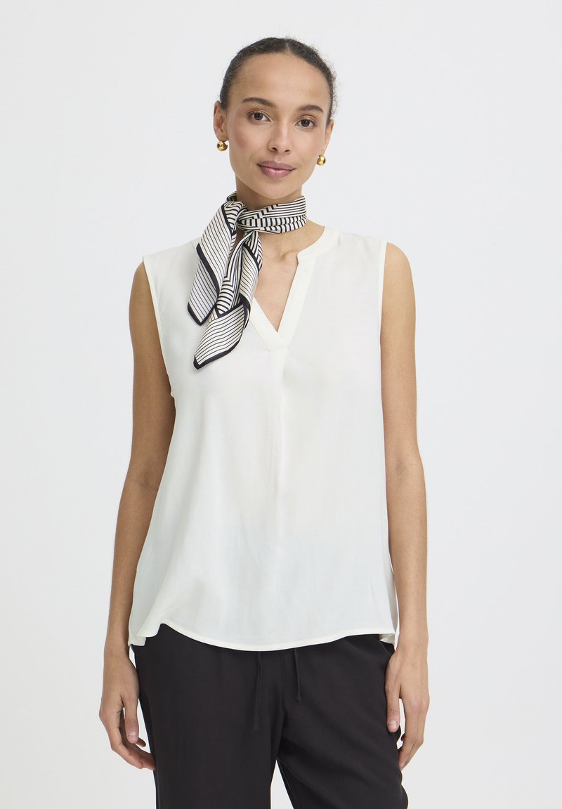 Woman wearing sleeveless white blouse, black trousers, striped black and white neck scarf, and gold hoop earrings, facing forward against white background.