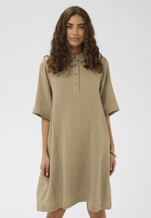 Młoda kobieta o kręconych włosach, ubrana w luźną beżową sukienkę do kolan z krótkimi rękawami i guzikami na przedniej listwie.