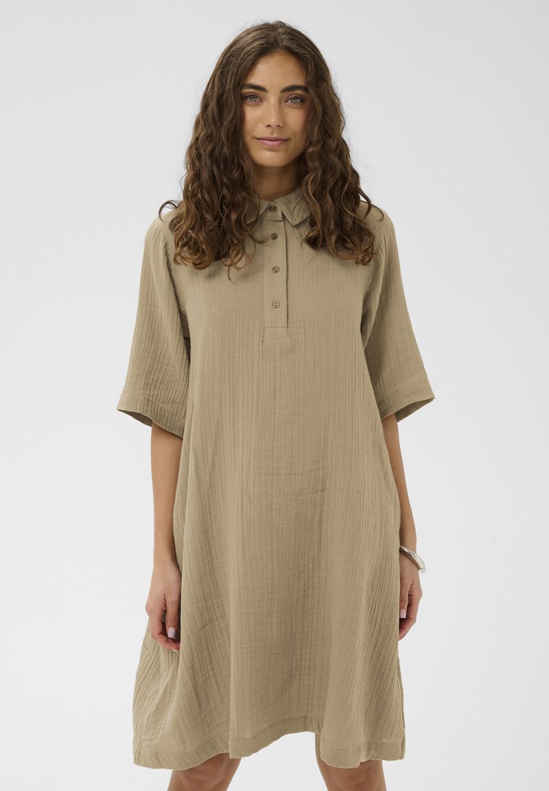 Jeune femme aux cheveux bouclés portant une robe beige ample, arrivant au genou, avec des manches courtes et des boutons sur la patte de boutonnage devant.
