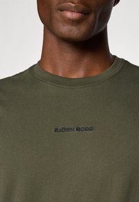 Detailný záber na osobu, ktorá má na sebe olivovo zelené tričko s vyšitým nápisom "BJÖRN BORG" v tmavej niťi na hrudi.