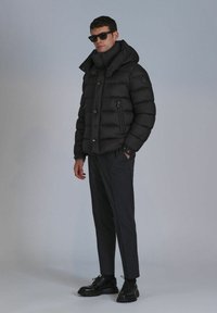 Black puffer jacket with a high collar and zippered pockets. Paired with dark trousers and black shoes. Model wearing sunglasses.