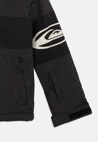 Crna jakna s uzorkom mreže, koja ima čvrsti crni panel na ramenu i bijeli logo na ruci. Uključuje džepove sa zatvaračem.