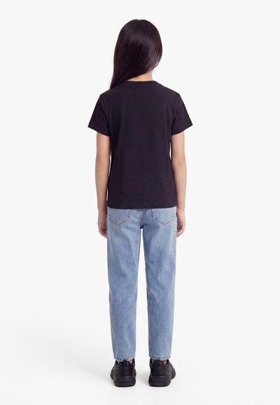 Young person with long dark hair wearing a black t-shirt, light blue jeans, and black sneakers, standing facing away on a white background.