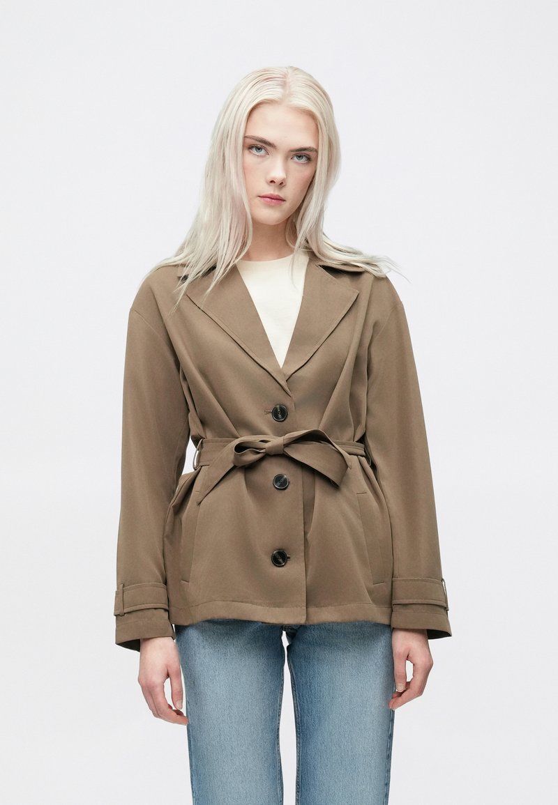 Braune Jacke mit Gürtel und großen Revers, ausgestattet mit drei schwarzen Knöpfen, Seitentaschen und einer glatten Stoffstruktur; kombiniert mit hellblauen Jeans.