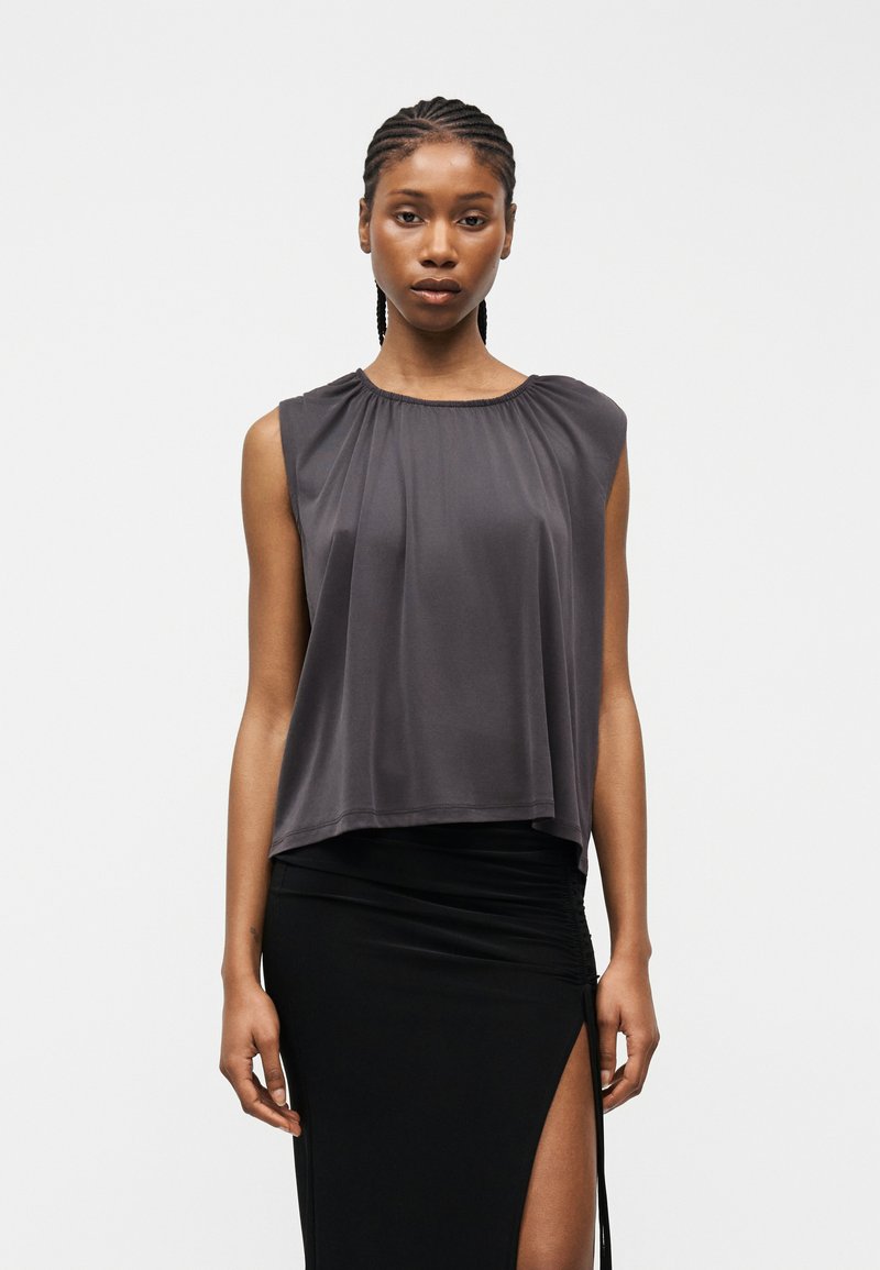 Femme aux cheveux tressés portant un haut gris sans manches et une jupe noire fendue, debout devant un fond clair uni.