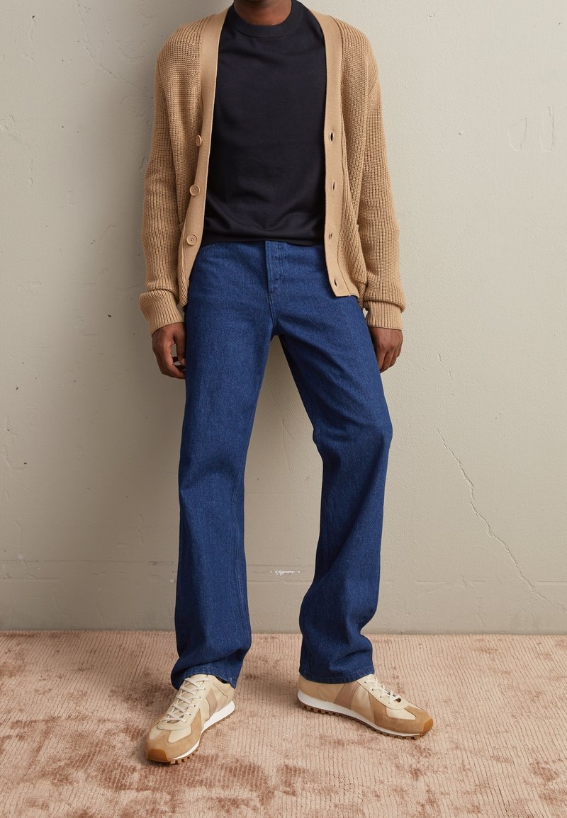 Homme portant un cardigan beige, un t-shirt noir, un jean bleu et des baskets beiges, debout contre un mur neutre et un sol moquetté.