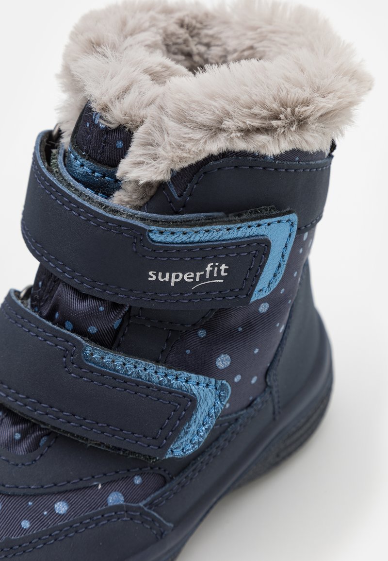 Marineblaues Kinderstiefel mit Kunstfellbesatz, Klettverschlüssen und einem Pünktchenmuster. Verfügt über einen glatten, glänzenden Akzent und eine strukturierte Sohle.