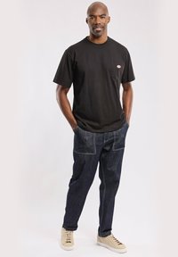 Schwarzes Baumwoll-T-Shirt mit Brusttasche, kombiniert mit dunklen Jeans aus Denim, die mehrere Nähte und hellen Stich haben. Beigefarbene Sneakers vervollständigen das Outfit.