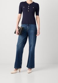 Granatowa prążkowana bluzka z krótkim rękawem i złotymi guzikami, w połączeniu z jasnoniebieskimi rozkloszowanymi dżinsami i cielistymi szpilkami, trzymająca czarną torebkę.