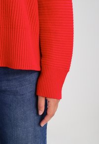 Red ribbed sweater with long sleeves and a wide cuff. Material appears knit with a textured finish. Worn over denim jeans.
