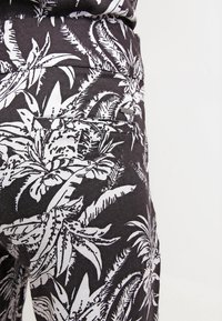 Zwarte broek met een wit tropisch bladrandpatroon, gemaakt van gladde stof, met een achterzak en een klein merketiketje nabij de tailleband.