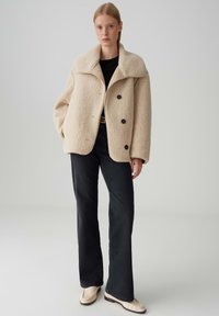 Vrouw staat in een beige shearling jas, zwart shirt, zwarte flared jeans, crèmekleurige loafers en een riem tegen een effen lichte achtergrond.