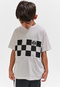 Boy wearing a white T-shirt with black and white checkered pattern across the chest, looking down with hands near his sides.
