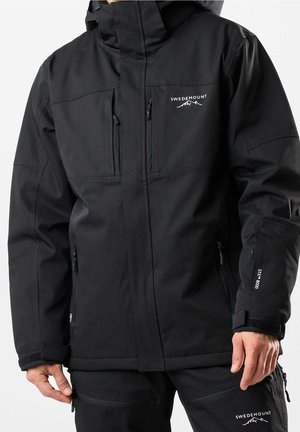 Volwassene draagt een zwarte Swedemount waterdichte jas met meerdere ritssluitingzakken en een bijpassende zwarte broek.