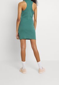 Sukienka bodycon w leśnej zieleni, bez rękawów z wysokim dekoltem. Modelka nosi jasnoróżowe sneakersy i białe skarpetki do kostek. Tekstura prążkowana przez całą długość.