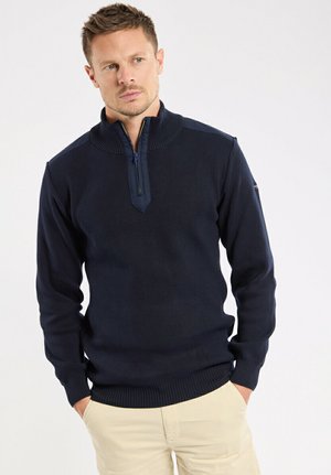 Homme portant un pull bleu marine à fermeture éclair partielle avec col et poignets côtelés, pantalon beige, debout les mains dans les poches devant un fond uni.