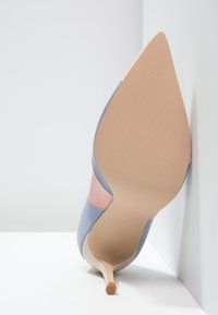 Ljusblå mocka högklackad sko med spetsig tå och rosa läderdetalj. Har en texturerad tan-sula. Anglad vy som fångar klacken.