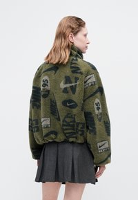 Oliivroheline fliisk jacket mustade graafiliste mustritega, kõrge krae ja elastse vöömisega. Kanda pleeditud halli seeliku peal.