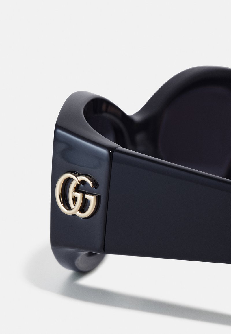 Schwarze Plastiksonnenbrille mit glänzender Oberfläche und einem goldenen GG-Logo-Akzent an der Seite. Das Design umfasst runde, dunkle Gläser.