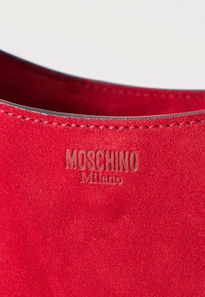 Κοντινό πλάνο κόκκινου σουέντ υφάσματος με ραμμένο άκρο και ανάγλυφο το εμπορικό όνομα "MOSCHINO Milano".