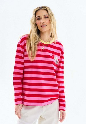 Kobieta o długich blond włosach, ubrana w czerwono-różową pasiastą bluzkę z długim rękawem i białe spodnie na jednolitym białym tle.