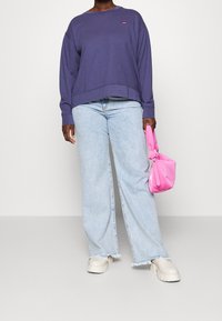 Persona che indossa jeans azzurri a gamba larga, scarpe chunky bianche, e una maglietta a maniche lunghe viola, mentre tiene una borsa rosa brillante su uno sfondo neutro.