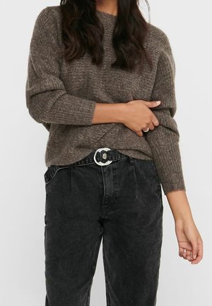 Frau trägt einen braunen Strickpullover mit langen Ärmeln und schwarze, hoch taillierte Jeans, die durch einen runden silbernen Schnallen-Gürtel gesichert sind.