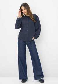 Marineblå bluse med knapfront og puffede ærmer, parret med brede, mørkeblå denimjeans og marineblå sko. Glat stoftekstur.
