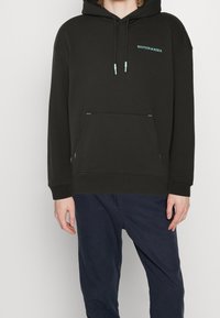 Sudadera negra con cordones ajustables y bolsillo frontal. Presenta acentos en turquesa en las costuras y el logo. Diseño suave y tipo pullover.