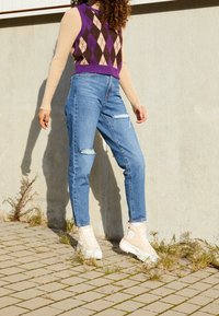 Chaleco de punto con patrón argyle en morado, marrón y beige; camiseta de manga larga color beige claro; jeans de mezclilla azul de talle alto con detalles desgastados; zapatillas beige.