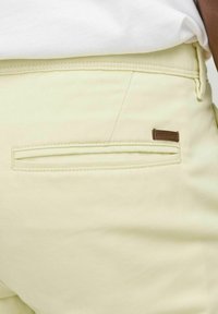 Pantalons jaune clair avec une texture lisse, comportant une poche arrière et une petite étiquette en cuir marron. La couture est appliquée uniformément.