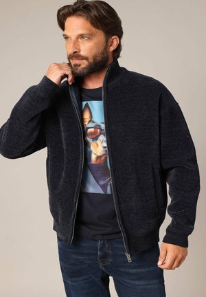Donkerblauwe, ritssluiting jack met een gestructureerd breipatroon, met zijzakken en een geribbelde rand. Draag het over een T-shirt met een hondenprint.