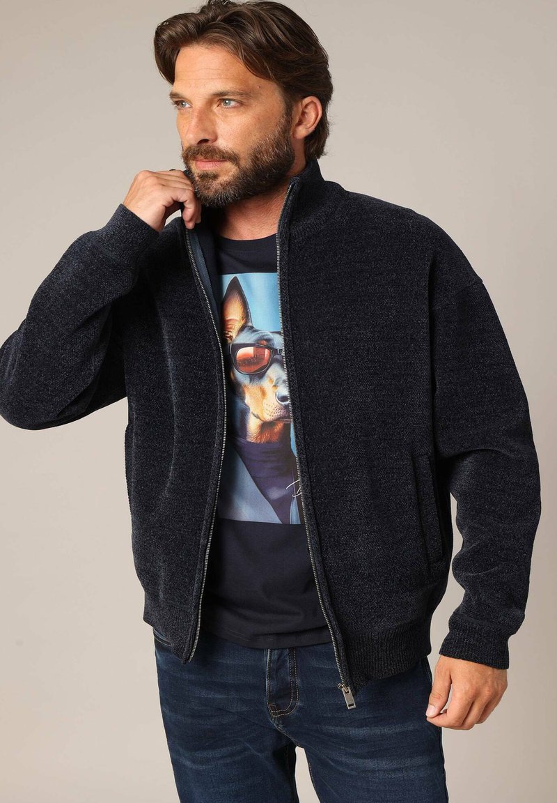 Veste zippée bleu marine foncé avec un design en tricot texturé, dotée de poches latérales et d'un ourlet côtelé. Portée par-dessus un t-shirt graphique avec un imprimé de chien.