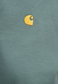 Βαμβακερό T-shirt σε απαλό teal με κίτρινο κεντημένο λογότυπο. Λείος υφασμάτινος χαρακτήρας, κοντά μανίκια και στρογγυλή λαιμόκοψη.