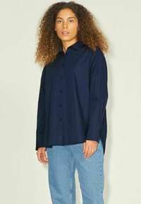 Camicia blu navy a maniche lunghe in un tessuto morbido, dotata di taschino sul petto e fessure ai lati per una maggiore ventilazione, abbinata a jeans azzurri.
