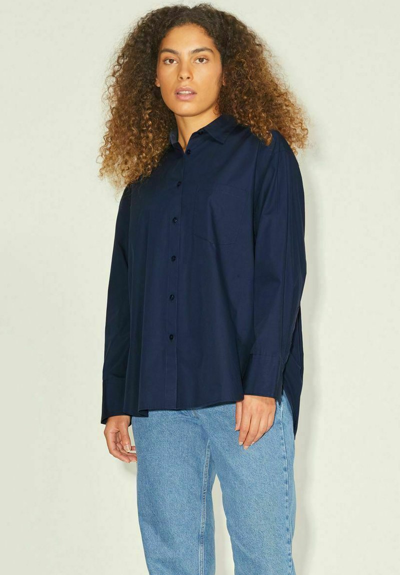 Camicia blu navy a maniche lunghe in un tessuto morbido, dotata di taschino sul petto e fessure ai lati per una maggiore ventilazione, abbinata a jeans azzurri.