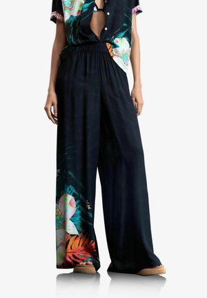 Pantalones negros de pierna ancha con estampado floral colorido cerca de los tobillos, combinados con una camisa oscura de flores de manga corta y zapatos sin cordones color canela.