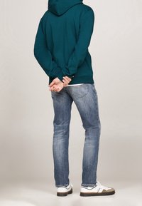 Sudadera color azul verdoso con capucha ajustable, llevada con jeans de un azul claro. Zapatillas blancas con detalles marrones. Vista desde atrás, con las manos en la espalda.