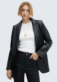 Černý blazer z umělé kůže se skládací klopou, uzávěrem na jeden knoflík a bočními kapsami, přeložený přes bílý upnutý top a černé kalhoty.