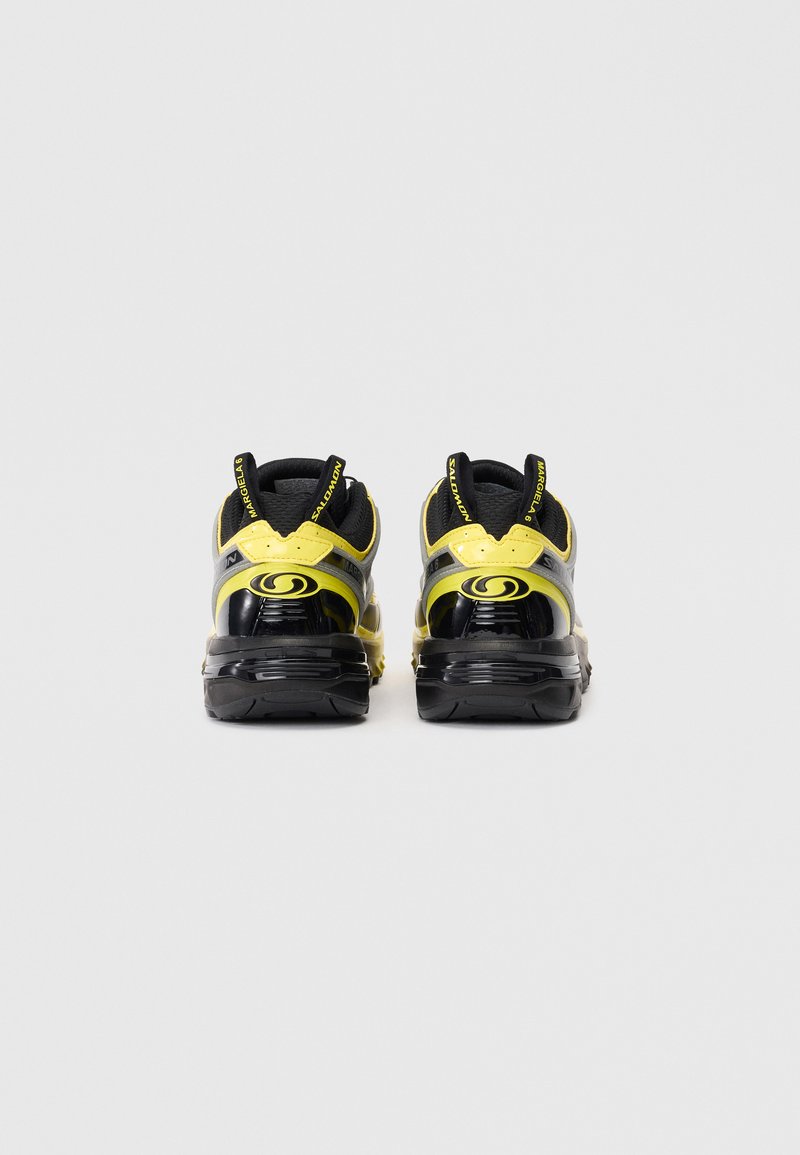 Des chaussures de sport noires et jaunes avec un design imposant, des surfaces lisses et texturées, des languettes de tirage au talon, et un logo distinctif à l'arrière.