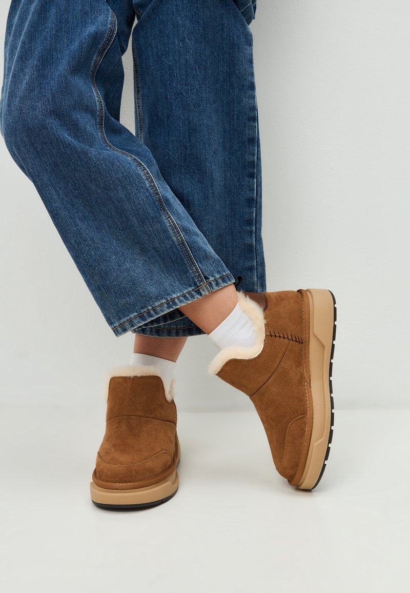 Persona che indossa jeans blu con risvolti, calzini bianchi e scarpe in suede marroni slip-on invernali con fodera in pelliccia leggera su un pavimento bianco.