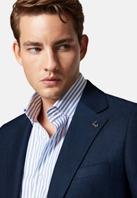 Granatowa marynarka z teksturowanym wykończeniem, noszona na jasnoniebieskiej koszuli w białe paski. Zauważalny detal: małe metaliczne logo na klapie marynarki.