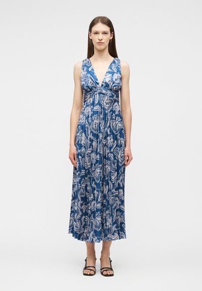 Femme portant une robe longue sans manches à motifs floraux bleus et blancs avec un décolleté en V profond et des sandales noires à lanières, debout devant un fond blanc.