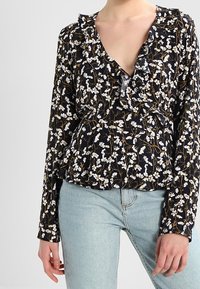 Blusa floreale con scollatura a V profonda, maniche lunghe, dettagli arricciati, sfondo nero, fiori bianchi e gialli, tessuto leggero.
