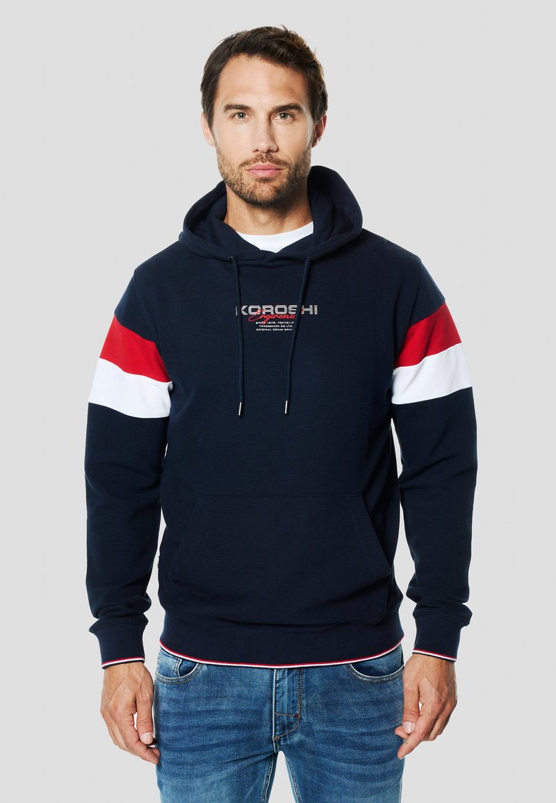 Tengerészkék kapucnis pulóver piros-fehér csíkos ujjakkal, kenguruzsebekkel és húzózsinóros kapucnival; texturált anyag díszítő logóval a mellkasnál.