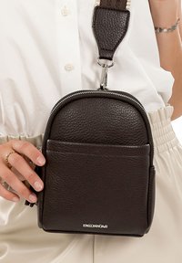 Small black textured leather crossbody bag with front pocket, silver zipper, and adjustable strap worn over white shirt and cream pants.