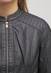 Černá kožená bunda s vertikálním zipem, prošívanými detaily na ramenech, kterou nosí osoba se světle hnědými vlasy a světlou pletí.