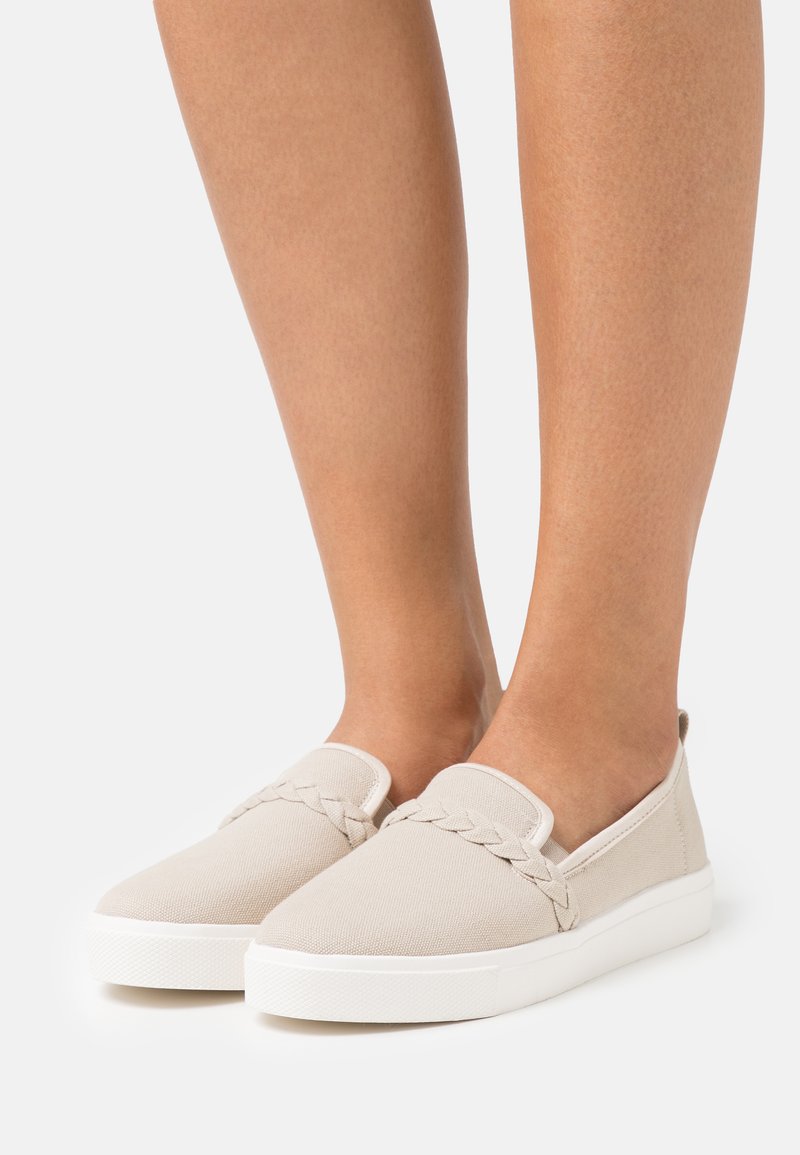 Beige Canvas Slip-on-Schuhe mit geflochtenem Akzent, ausgestattet mit einer flachen weißen Gummisohle und glatter Textur, die ein minimalistisches Design präsentiert.